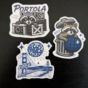 May include: Set of three stickers featuring raccoon illustrations. One sticker shows a raccoon behind a crate with the word "Portola". Another shows a raccoon in a trash can with a globe and the number "2025". The third sticker depicts the Golden Gate Bridge and a disco ball with the number "2025".