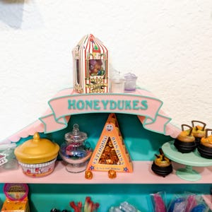 May include: A pink and teal display shelf labeled HONEYDUKES featuring miniature candy boxes and jars. Items include a striped Every Flavour Beans box, a yellow lidded container, a glass jar, a triangular Exploding Bonbons box, and small black cauldrons.