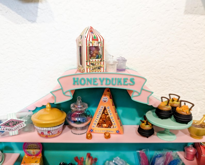 May include: A pink and teal display shelf labeled HONEYDUKES featuring miniature candy boxes and jars. Items include a striped Every Flavour Beans box, a yellow lidded container, a glass jar, a triangular Exploding Bonbons box, and small black cauldrons.