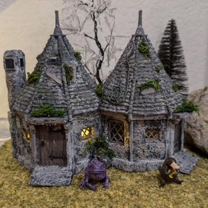 Może przedstawiać: Miniaturowy kamienny domek z ostrym dachem, małym kominem i drewnianymi drzwiami. Domek jest ozdobiony mchem i winoroślą, a przed nim stoją dwie małe figurki. Domek stoi na łóżku z brązowej trawy.