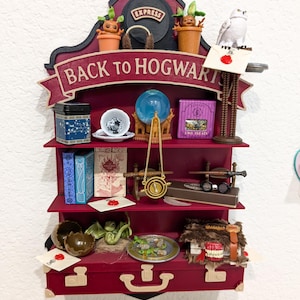 May include: A burgundy shelf unit with a train-shaped top, displaying miniature figurines and themed items. The words "BACK TO HOGWARTS" are prominently displayed. Items include a snowy owl, a teacup, a blue orb, and a miniature suitcase.