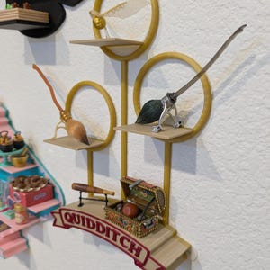 May include: A decorative Quidditch-themed display featuring a golden snitch, brooms, and a treasure chest. The display includes the word "Quidditch" on a banner. The items are arranged on shelves with a gold circular design.