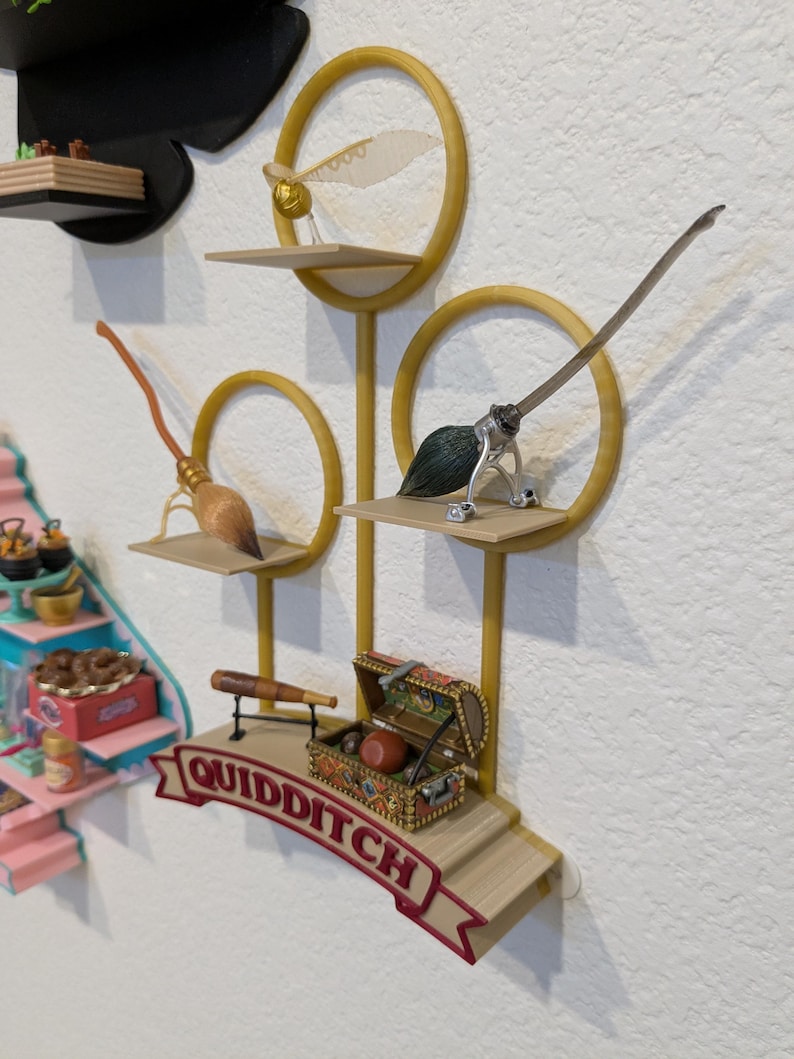 May include: A decorative Quidditch-themed display featuring a golden snitch, brooms, and a treasure chest. The display includes the word "Quidditch" on a banner. The items are arranged on shelves with a gold circular design.