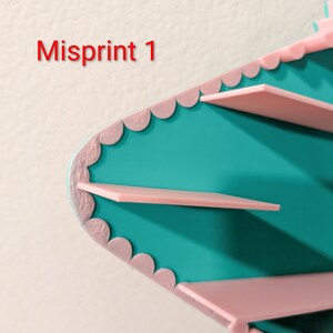 May include: A teal shelf unit with light pink scalloped edges and horizontal pink shelves. The text Misprint 1 is visible in red on the left side. The structure features a pointed, angular design with a smooth finish on the teal surfaces.