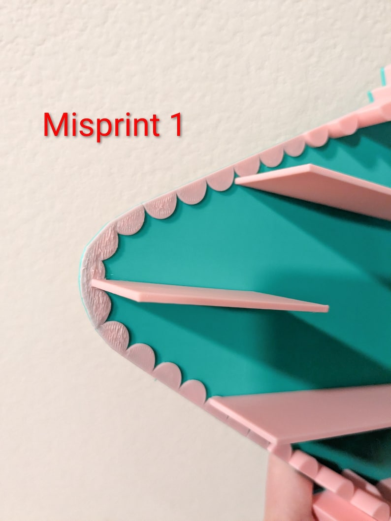 May include: A teal shelf unit with light pink scalloped edges and horizontal pink shelves. The text Misprint 1 is visible in red on the left side. The structure features a pointed, angular design with a smooth finish on the teal surfaces.