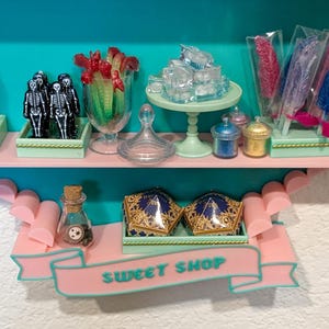 May include: A miniature sweet shop display featuring two shelves with various candy-themed items, including skeleton figures, rock candy sticks, small jars, and two gold-patterned boxes. A pink banner on the lower shelf displays the text SWEET SHOP.