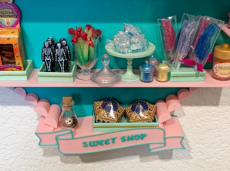 May include: A miniature sweet shop display featuring two shelves with various candy-themed items, including skeleton figures, rock candy sticks, small jars, and two gold-patterned boxes. A pink banner on the lower shelf displays the text SWEET SHOP.