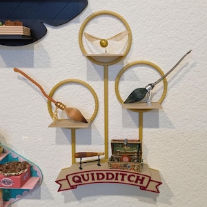 May include: A decorative wall display featuring a golden snitch, two miniature broomsticks, and a treasure chest. The display includes the word "QUIDDITCH" on a banner. The items are arranged on a gold-colored frame with circular accents.