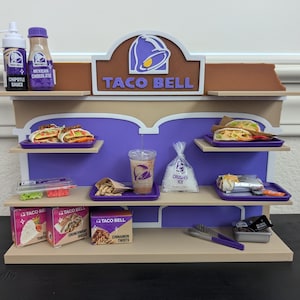 Puede incluir: Una maqueta de Taco Bell con tacos, burritos y cinnamon twists. La exhibición incluye botellas de salsa Chipotle y chocolate mexicano, un vaso de hielo y cajas. El logotipo de Taco Bell es visible. La exhibición es de madera y pintada.