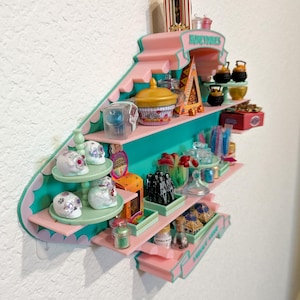 May include: A wall-mounted display shelf in pink and teal featuring the text Honeydukes. The shelf holds miniature candy-themed items including white skull figurines, small jars, colorful boxes, and tiny cauldrons arranged on multiple tiers.