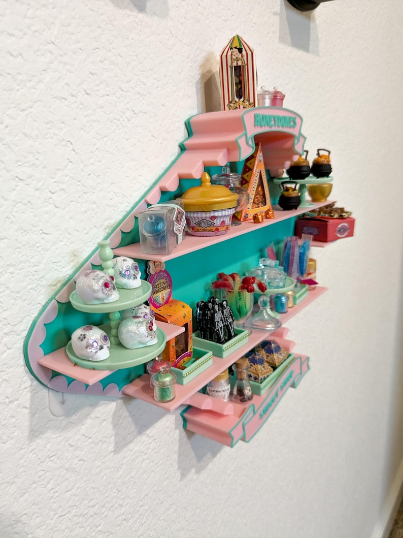 May include: A wall-mounted display shelf in pink and teal featuring the text Honeydukes. The shelf holds miniature candy-themed items including white skull figurines, small jars, colorful boxes, and tiny cauldrons arranged on multiple tiers.