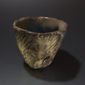 Puede incluir: Una taza pequeña de cerámica marrón con un diseño texturizado blanco y marrón. La taza tiene un borde ligeramente acampanado y una base redondeada.