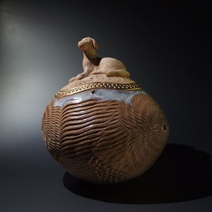 以下が含まれることがあります： テクスチャのある茶色の外側と、小さな羊の彫刻が施された蓋が付いたセラミック製の壺。蓋は金色の帯と小さな金色のアクセントで飾られています。