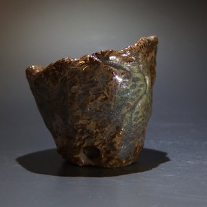 Puede incluir: Una pequeña maceta de cerámica marrón con una superficie rugosa y texturizada. La maceta tiene una forma única e irregular y un esmalte ligeramente verde.