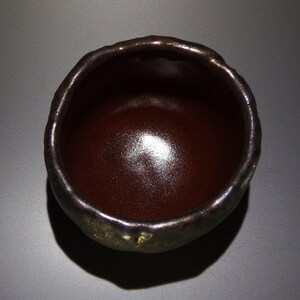 May include: A dark brown ceramic teacup with a slightly uneven rim. The cup has a glossy finish and a subtle sheen.