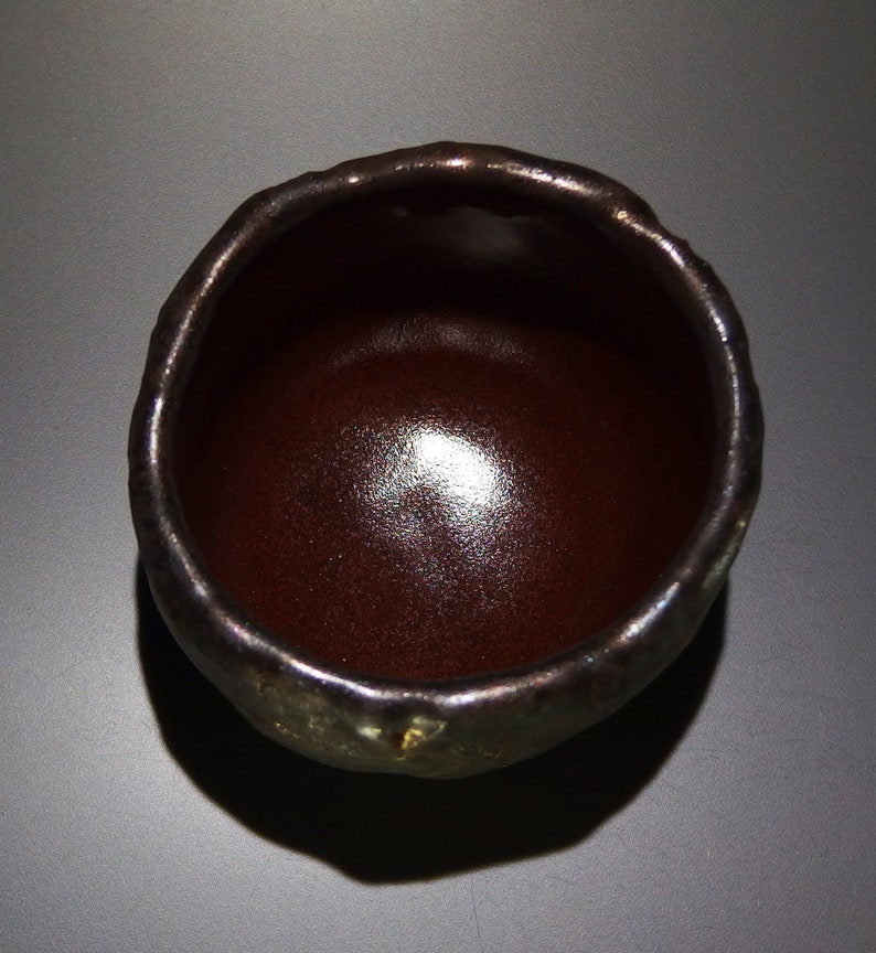 May include: A dark brown ceramic teacup with a slightly uneven rim. The cup has a glossy finish and a subtle sheen.