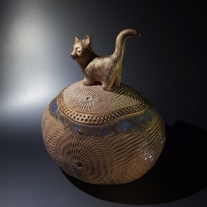 以下が含まれることがあります： 金色の猫の置物が置かれた、卵型のセラミック製の容器。容器は、青と金の模様が施された質感のある表面をしています。