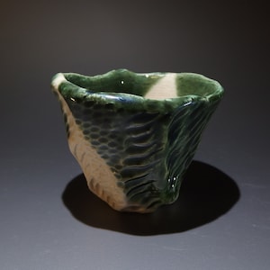 Puede incluir: Una taza pequeña de cerámica verde y blanca con un diseño texturizado en forma de onda. La taza está hecha a mano y tiene una forma irregular.