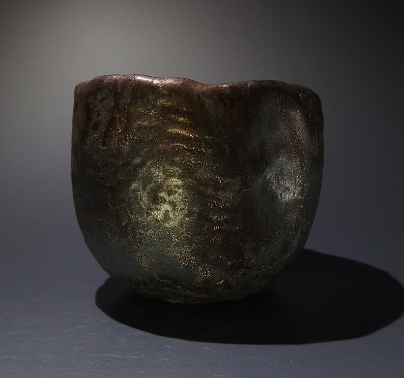 May include: A dark brown ceramic bowl with a textured, uneven surface. The bowl has a metallic sheen with hints of gold and green.