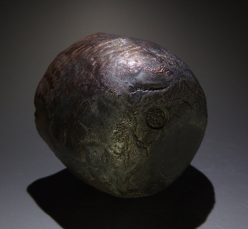 May include: A dark gray ceramic pot with a textured surface and a circular mark on the side.