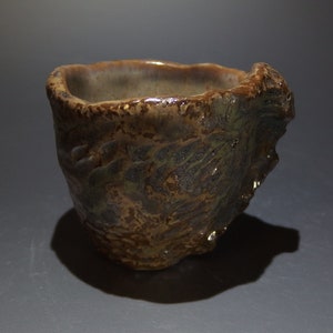 Puede incluir: Una taza pequeña de cerámica marrón con una superficie rugosa y texturizada. La taza tiene un borde ancho y redondeado y una base ligeramente irregular.