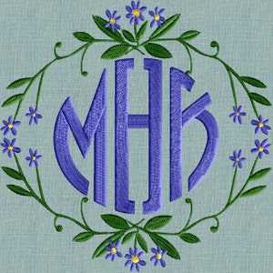 Op de afbeelding: Een monogram met de letters "MHB" in een blauw lettertype, omringd door een krans van groene bladeren en paarse bloemen.