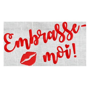 May include: Red embroidered text on a white fabric background that says "Embrasse-moi!" with a red embroidered heart and a red embroidered lip print.