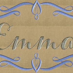 Puede incluir: Un fondo de tela beige con un diseño bordado en azul y gris. El diseño presenta un borde decorativo con un corazón estilizado en el centro y el nombre "Emma" en una fuente cursiva.