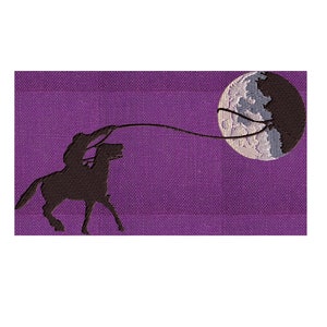 May include: A silhouette of a person riding a horse and lassoing the moon. The moon is a white and gray crescent moon with a dark gray background. The background is a purple textured fabric.