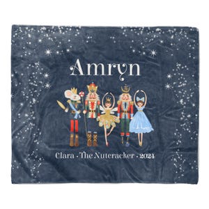 May include: A blue fleece blanket with a starry night design. The blanket features a personalized name "Amryn" and a scene from the Nutcracker ballet. The text "Clara. The Nutcracker. 2024" is printed at the bottom.