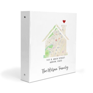May include: A white three-ring binder featuring a house-shaped map graphic with a red heart icon above the chimney. The text 145 S. MAIN STREET AKRON, OHIO is printed below the map, followed by the script text The Wilson Family at the bottom.