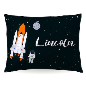 May include: A black pillow with a space theme. It features a white space shuttle with orange flames, a white astronaut, a grey moon, and white stars. The name "Lincoln" is written in white cursive lettering.