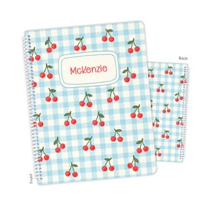 May include: A spiral-bound notebook featuring a light blue and white gingham pattern with a repeating cherry print. The front cover includes a central white rectangular label with a blue border containing the name Mckenzie in red text.