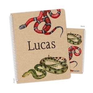 May include: A brown notebook with a spiral binding features a red and black snake on the front cover and a green and black snake on the back cover. The notebook has the name "Lucas" printed on the front cover.