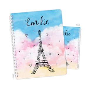 May include: A spiral notebook with a watercolor background of pink, blue, and yellow clouds. The Eiffel Tower is in the center of the notebook with hearts floating around it. The front cover says "Emilie".