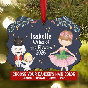 May include: A dark blue, scalloped-edge ornament featuring a nutcracker, a small child in a white dress, and a ballerina in a pink tutu. The text reads Isabelle Waltz of the Flowers 2026. A red ribbon is attached to the top for hanging.
