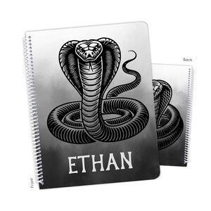 May include: A spiral-bound notebook featuring a black and white illustration of a cobra snake with the name "ETHAN" in bold white letters. The background has a gradient effect, transitioning from white to dark gray.