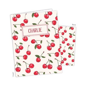 May include: A notebook and a smaller notepad feature a cream background with a pattern of red cherries and green leaves. The notebook has the name "CHARLIE" in a rectangular frame. The notepad's back is also visible.