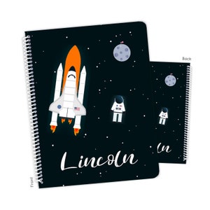 May include: A black notebook with a space theme. The front cover features a white space shuttle with orange flames, a moon, and a white astronaut. The back cover features a moon and an astronaut. The notebook has a white spiral binding and the name "Lincoln" is written in white cursive on the front cover.