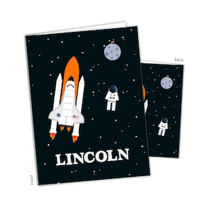 May include: A greeting card featuring a space theme with a black background filled with white stars. The design includes an orange and white space shuttle, an astronaut, a gray moon with a flag, and the name LINCOLN printed in white capital letters.