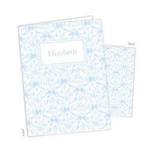 May include: A light blue and white stationery set featuring a floral and bow pattern. The front of the notebook is labeled "Elizabeth" in a white rectangular box. The back of the notebook is also visible.