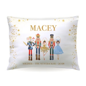 May include: A white pillow with gold glitter stars and the name "MACEY" in gold lettering. The pillow features a colorful illustration of a Nutcracker, a ballerina, a soldier, and a mouse. The text "SOLDIER - THE NUTCRACKER - 2018" is printed below the illustration.