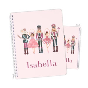 May include: Pink spiral notebook with a front cover featuring a watercolor illustration of a Nutcracker and a ballerina. The illustration includes a mouse in a crown and a second ballerina. The name "Isabella" is printed in purple at the bottom of the cover.
