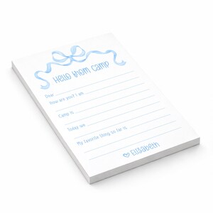 May include: White notepad with light blue watercolor bow and text "Hello from camp". The notepad has lines for writing and the name "Elizabeth" at the bottom. A cute stationery item.