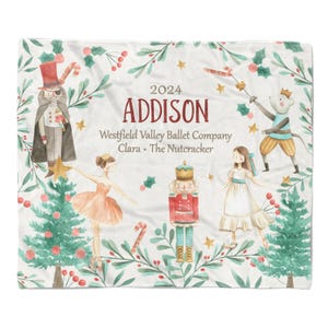 May include: A white fleece blanket with a watercolor design featuring characters from the Nutcracker ballet. The blanket has a green, red, and gold color scheme and includes the text "2024 ADDISON Westfield Valley Ballet Company Clara - The Nutcracker".