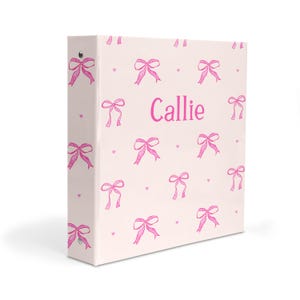 May include: A pink binder with a white background and a pattern of pink bows. The binder has the name "Callie" printed on the front.