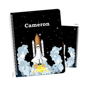 May include: A spiral-bound notebook with a black cover featuring a white rocket ship blasting off. The name "Cameron" is printed in white. The background is a starry night sky with white stars and blue clouds.