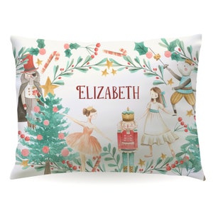 May include: A white pillow with a watercolor design featuring a ballerina, a nutcracker, a mouse king, and a Christmas tree. The pillow has a green wreath border with red berries and gold stars. The text "ELIZABETH" is printed in red on the pillow.