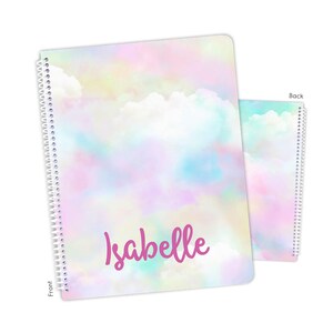 May include: A spiral-bound notebook featuring a pastel cloud pattern in shades of pink, blue, and yellow. The front cover displays the name Isabelle in a magenta script font. The image shows both the front and back covers of the notebook.