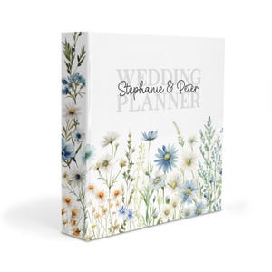 May include: A white three-ring binder featuring a watercolor illustration of blue and white wildflowers along the bottom and spine. The text "WEDDING PLANNER" is printed in gray, with the names "Stephanie & Peter" in black script above.